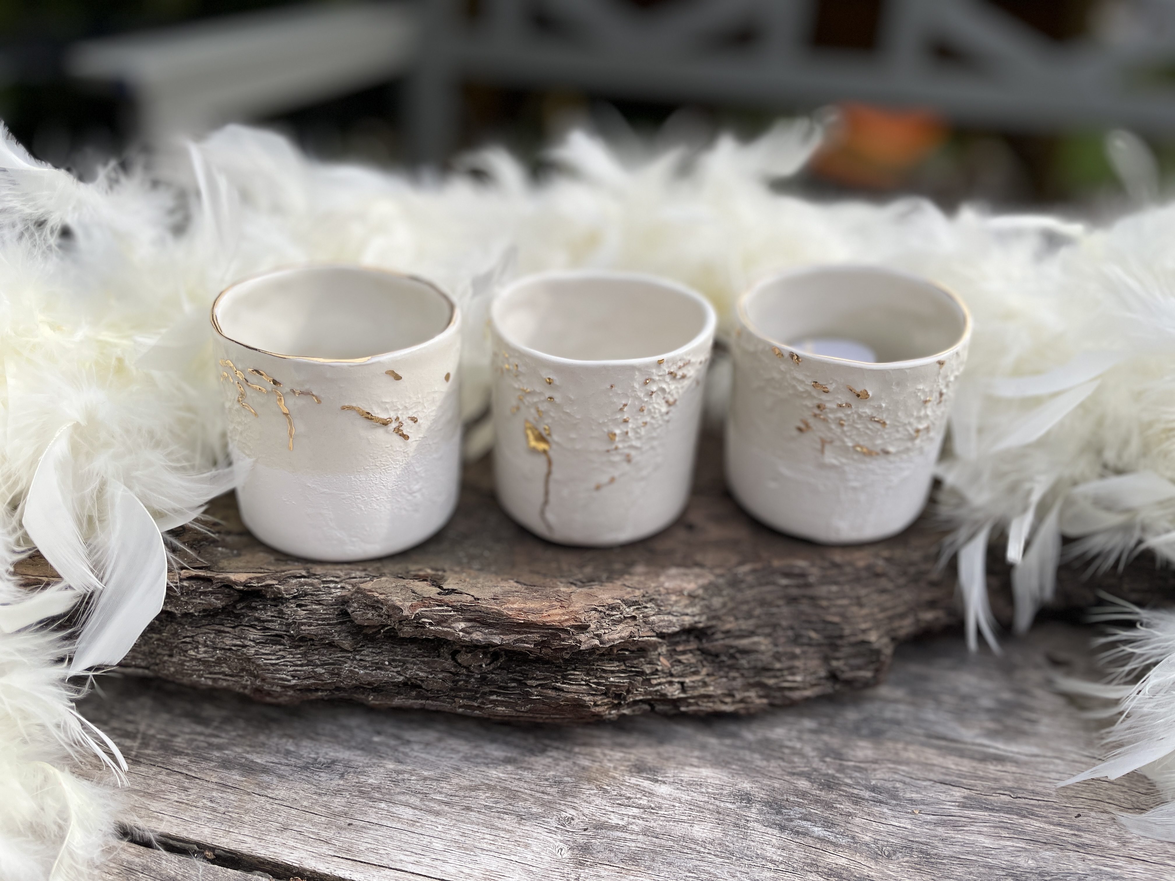 Drei weiße Keramikbecher mit Goldkintsugi auf Treibholz, umgeben von Federn.